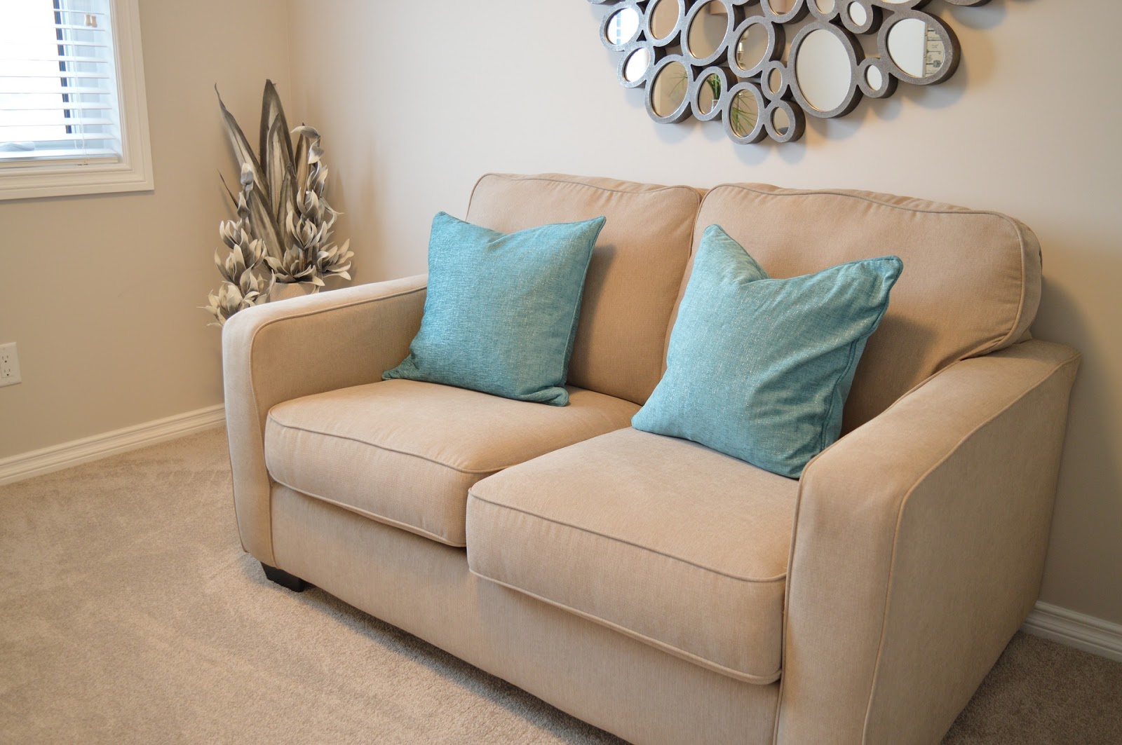 Foto de um sofá bege com almofadas azuis em uma sala. Vemos um quadro, planta e janela ao lado. Imagem ilustrativa para texto mistura para limpar sofá.